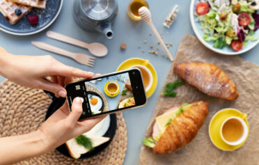 weibliche Hände halten Smartphone und fotographieren Frühstück mit Croissants und Gemüse