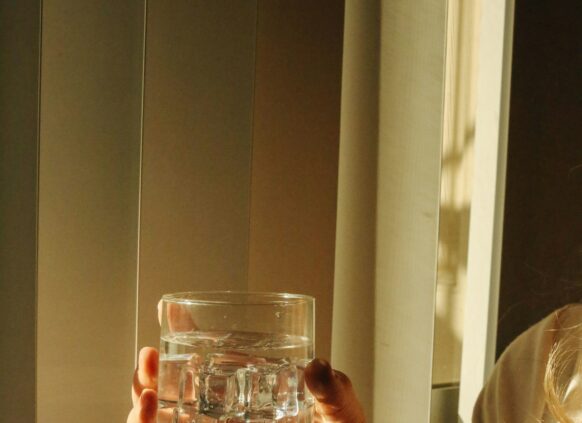 Hand hält ein Glas Wasser vor einem Fenster in warmen Licht.