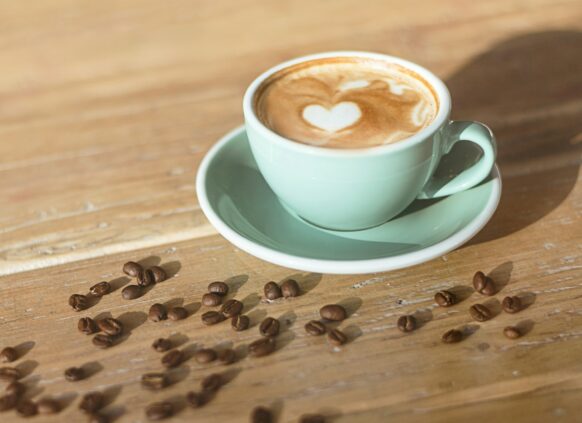 Kaffee in mint farbener Tasse auf einer Holzplatte, auf der Kaffeebohnen verteilt sind.