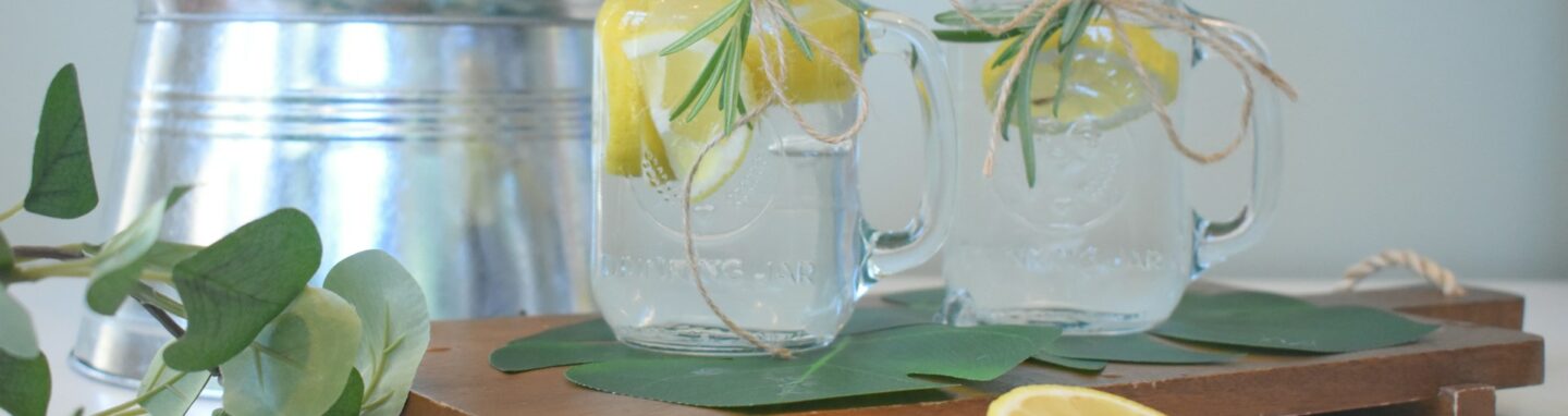 Zwei Gläser befüllt mit Wasser und Zitronen auf einem Holzbrett angerichtet.