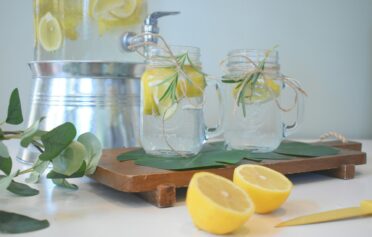 Zwei Gläser befüllt mit Wasser und Zitronen auf einem Holzbrett angerichtet.