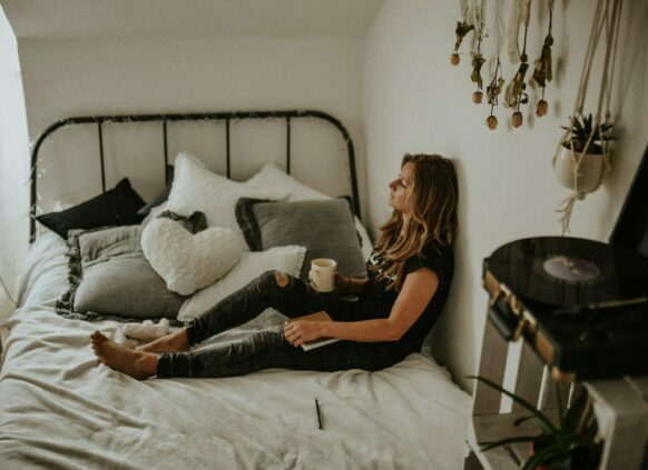 Frau sitzt mit einr Tasse auf einem Bett und guckt gerade aus.