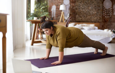 Attractive barefoot young overweight female doing plank on yoga mat while training indoors, watching online video via laptop. Sports, well being, technology and active healthy lifestyle concept