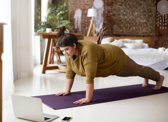 Attractive barefoot young overweight female doing plank on yoga mat while training indoors, watching online video via laptop. Sports, well being, technology and active healthy lifestyle concept