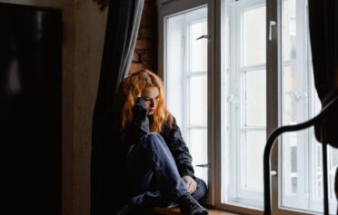 Junge Frau sitzt auf einer Fensterbank vor einem Fenster ud schaut traurig.