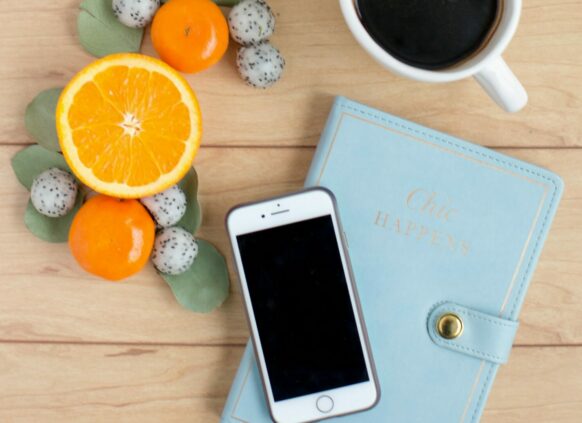 Auf einer Tischplatte liegt ein blaues Notizbuch, darauf steht eine Tasse Kaffe und ein Handy. Daneben liegt Obst.