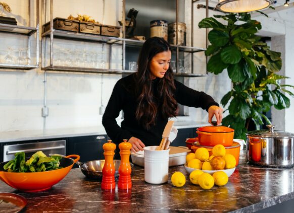 Frau mit braunen langen Haaren in schwarzen Oberteil steht in der Küche. Um sie herum liegen Orangen und ein orangener Kochtopf