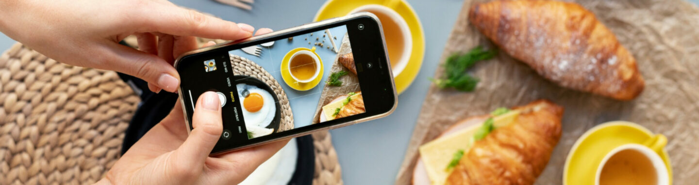 weibliche Hände halten Smartphone und fotographieren Frühstück mit Croissants und Gemüse