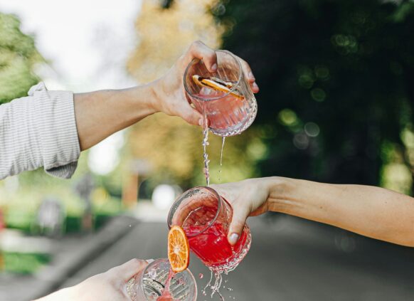 Vier Hände halten verschiedene Gläser mit roter Limonde und schütten diese aus, sodass die Limonde über die anderen Gläser darunter fließt.