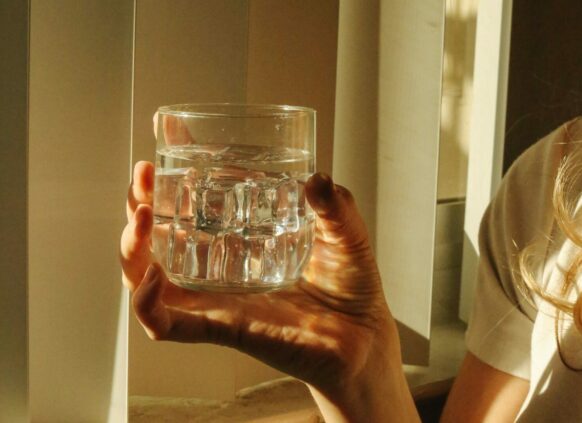 Hand hält ein Glas Wasser vor einem Fenster in warmen Licht.