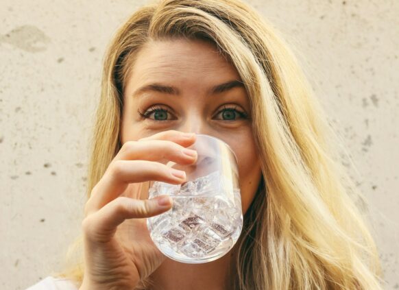 Frau mit blonden Haaren trinkt ein Glas Wasser und guckt in die Kamera.
