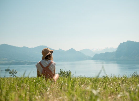Frau sitzt entspannt im Feld neben einem See, die Sonne scheint.