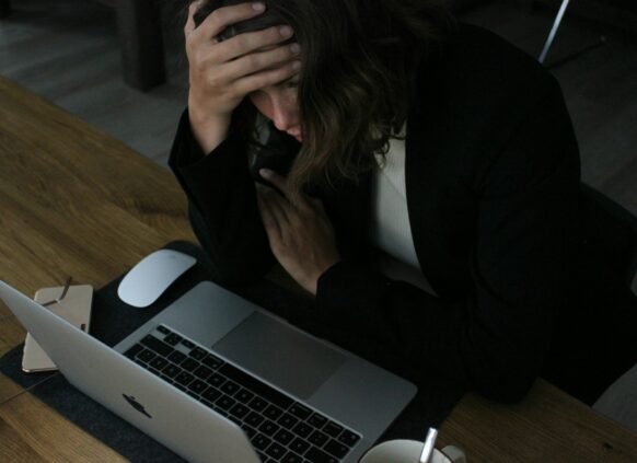 Eine gesresste Frau sitzt vor ihrem Laptop und fässt sich an die Stirn.
