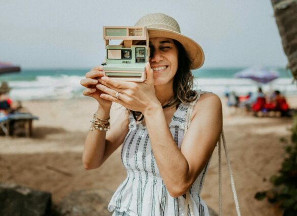 Eine Frau hält eine Sofortbildkamera vor ihrem Gesicht und macht ein Foto am Strand.