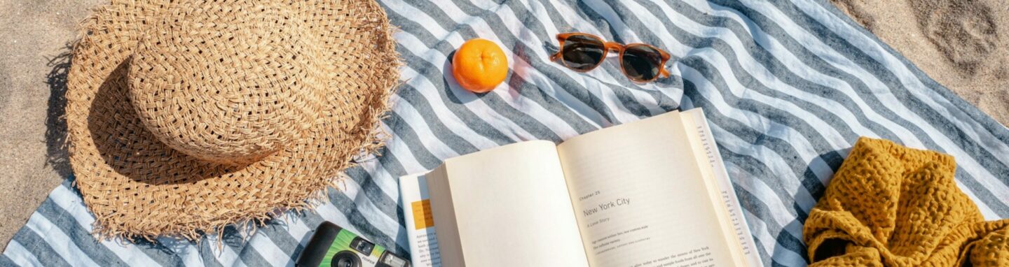 Auf einem gestreiften Handtuch am Strand liegen ein Sonnenhut, eine Sonnenbrille und ein Buch.