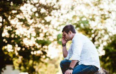Ein Mann im weißen Hemd sitzt auf einer Parkbank und fässt sich gestresst an die Stirn